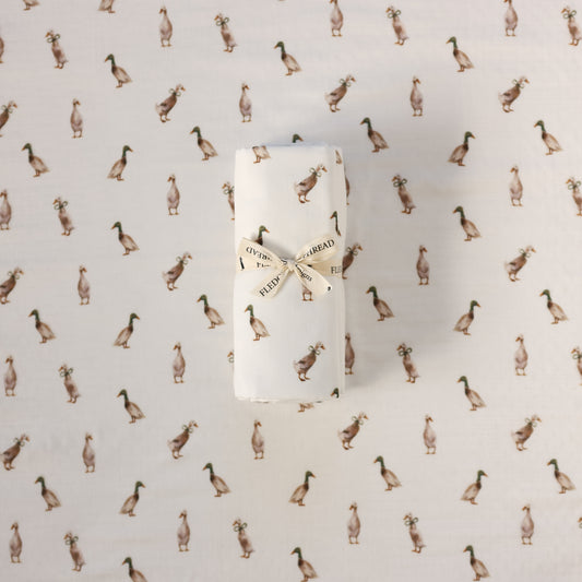 A rolled white swaddle, tied with a cream ribbon printed with the words 'FLEDGE AND THREAD', placed on a white fabric background patterned with small brown ducks.