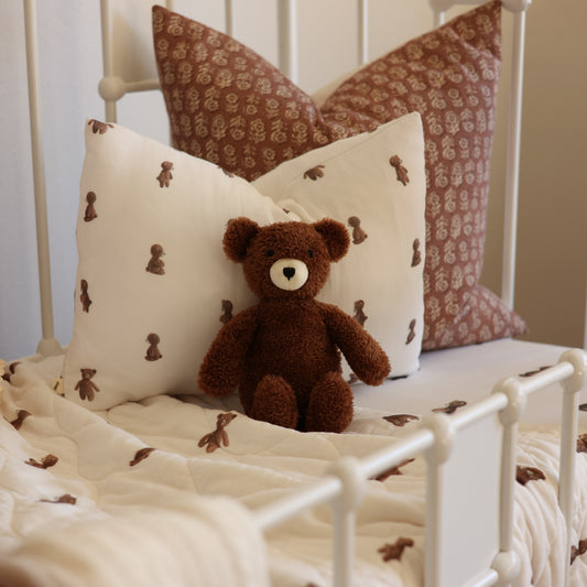 A cozy bed styled with a brown plush teddy bear sitting on a cream quilt and matching pillows, including two patterned brown pillows with teddy bear motifs, set in a white metal bed frame.