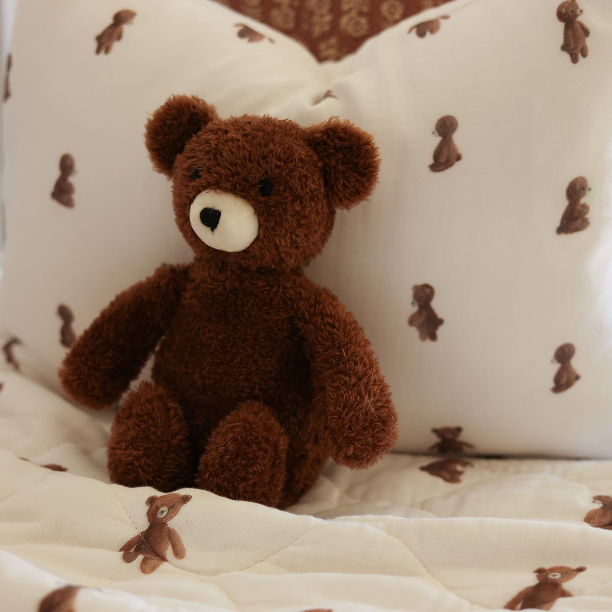 A brown plush teddy bear sitting on a quilted cream bedspread with matching pillows printed with small teddy bear motifs.