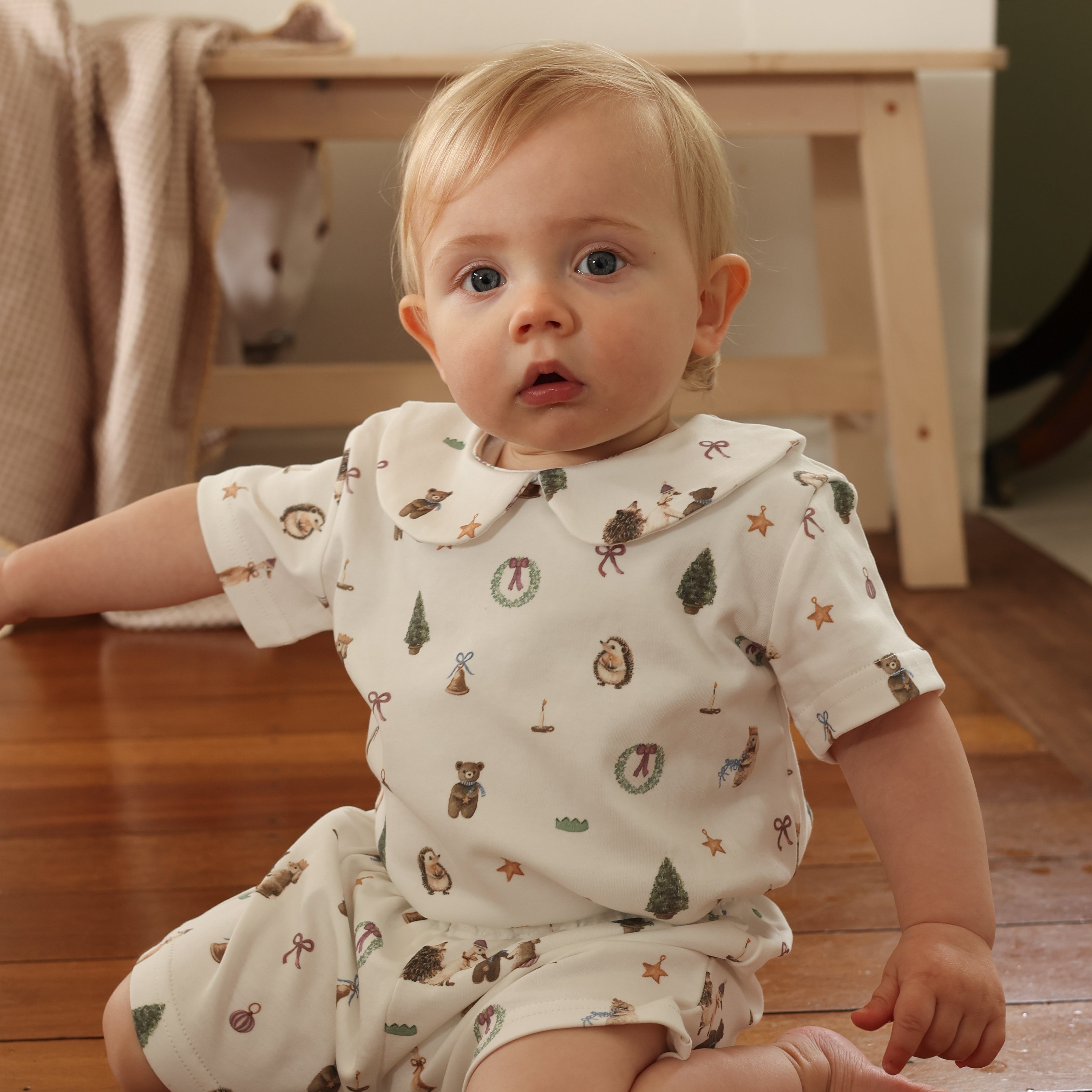Baby wearing a white outfit with animal patterns sitting on a wooden floor.