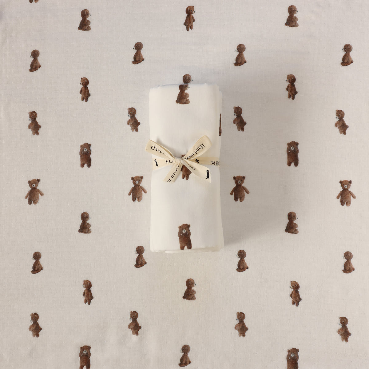 A rolled white fabric item, likely a baby blanket or swaddle, tied with a cream ribbon printed with teddy bears, placed on top of a matching white fabric background patterned with small brown teddy bears.
