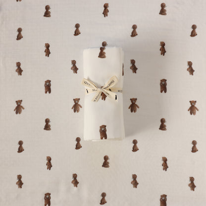 A rolled white fabric item, likely a baby blanket or swaddle, tied with a cream ribbon printed with teddy bears, placed on top of a matching white fabric background patterned with small brown teddy bears.