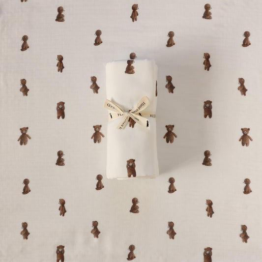 A rolled white fabric item, likely a baby blanket or swaddle, tied with a cream ribbon printed with teddy bears, placed on top of a matching white fabric background patterned with small brown teddy bears.