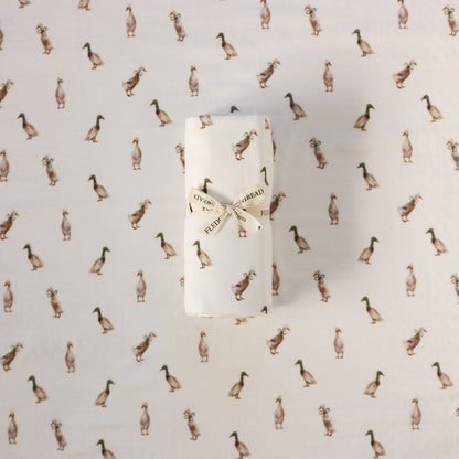 A rolled white swaddle, tied with a cream ribbon printed with the words 'FLEDGE AND THREAD', placed on a white fabric background patterned with small brown ducks.