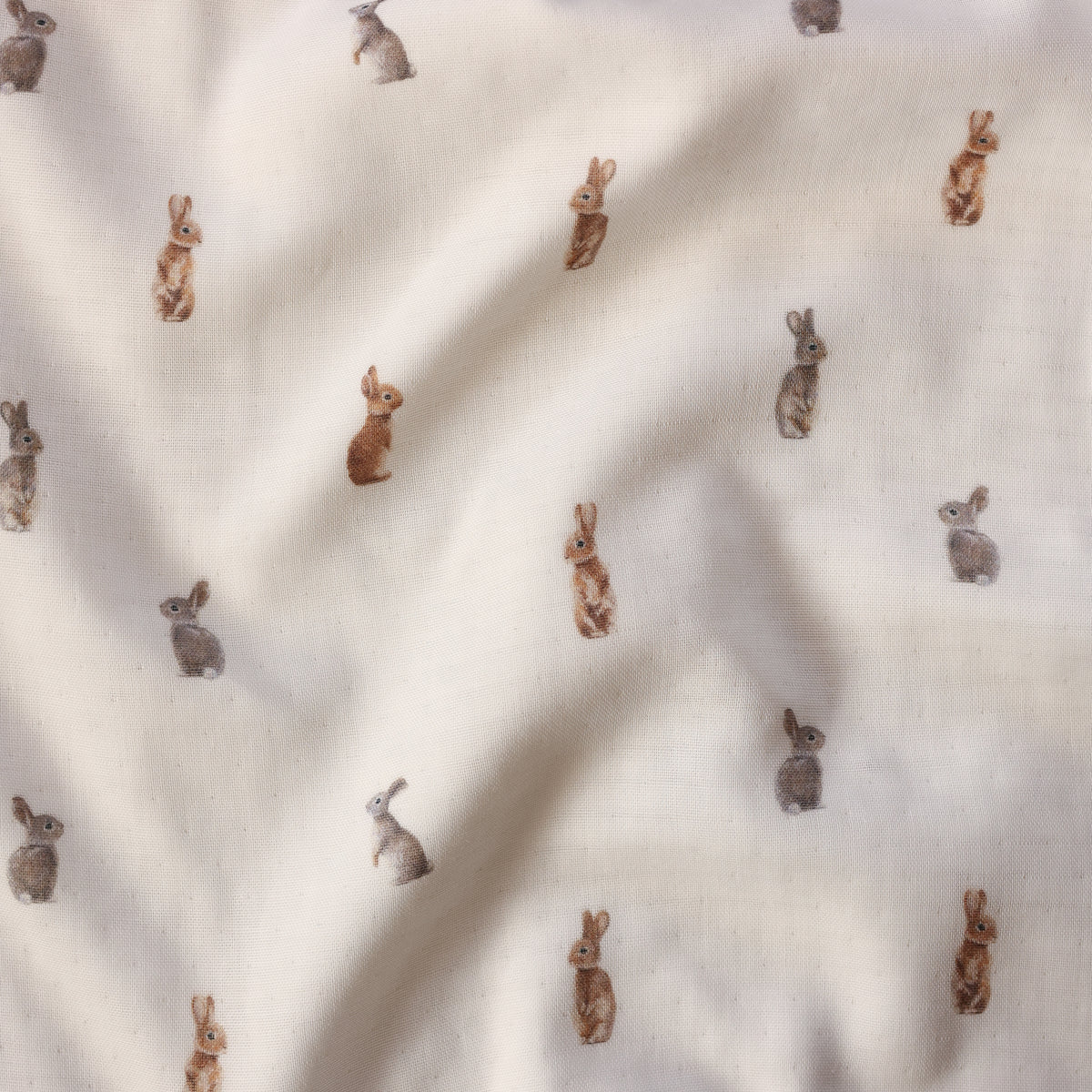 A close-up of a cream-colored fabric with a repeating pattern of small rabbits in brown, tan, and gray tones.