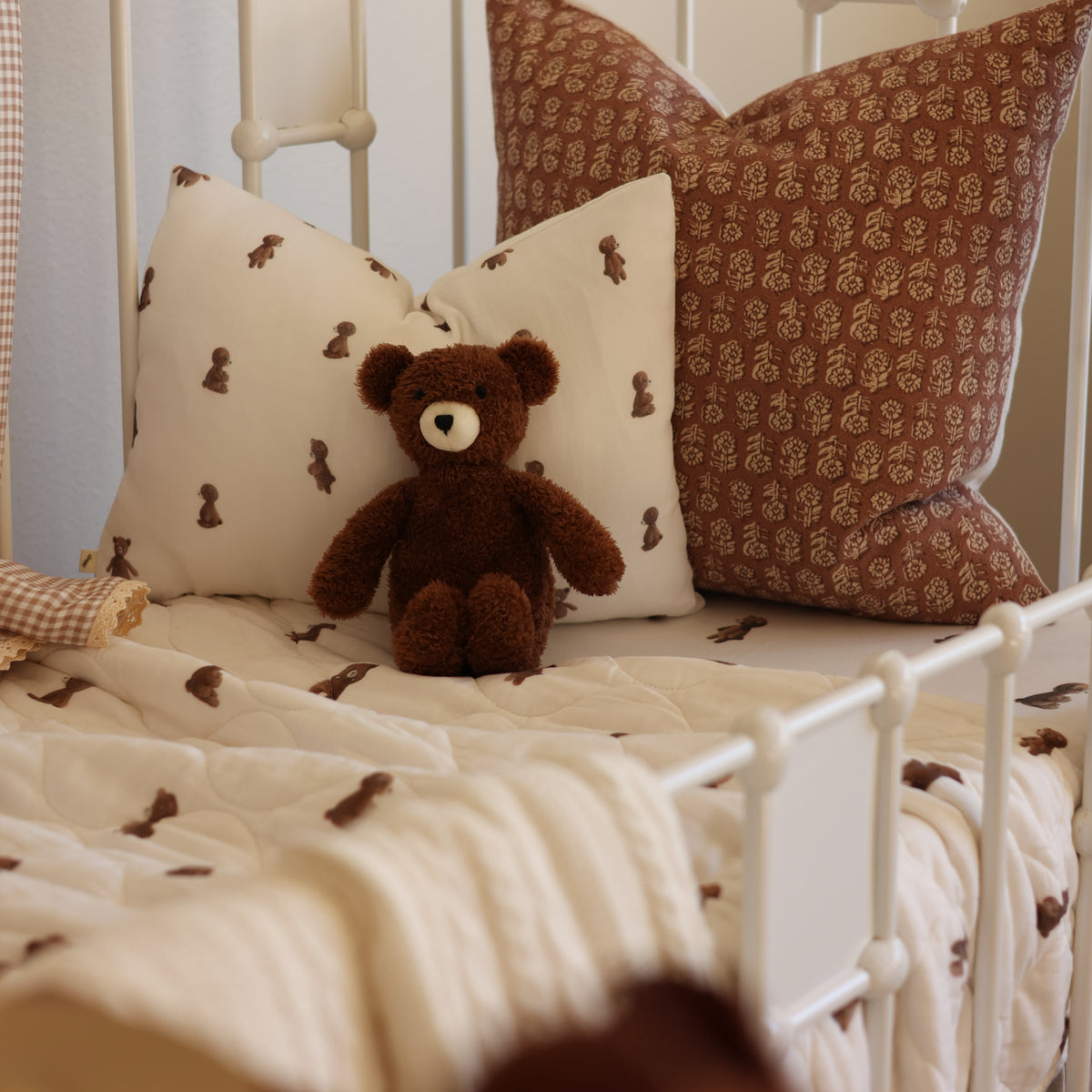 A cozy bed setup with cream and brown patterned pillows, a matching quilted blanket, and a small brown teddy bear sitting in the center. The bed has a white metal frame and is styled in a soft, warm bedroom setting.