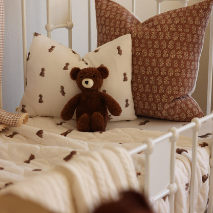 A cozy bed setup with cream and brown patterned pillows, a matching quilted blanket, and a small brown teddy bear sitting in the center. The bed has a white metal frame and is styled in a soft, warm bedroom setting.