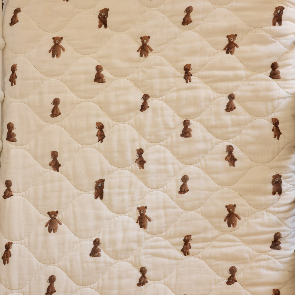 A close-up of a quilted fabric surface in a warm cream or off-white tone, patterned with small brown teddy bears repeated across the material.