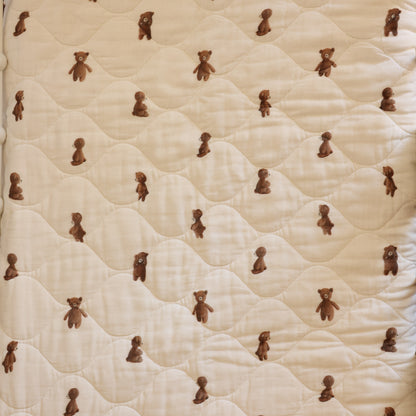 A close-up of a quilted fabric surface in a warm cream or off-white tone, patterned with small brown teddy bears repeated across the material.