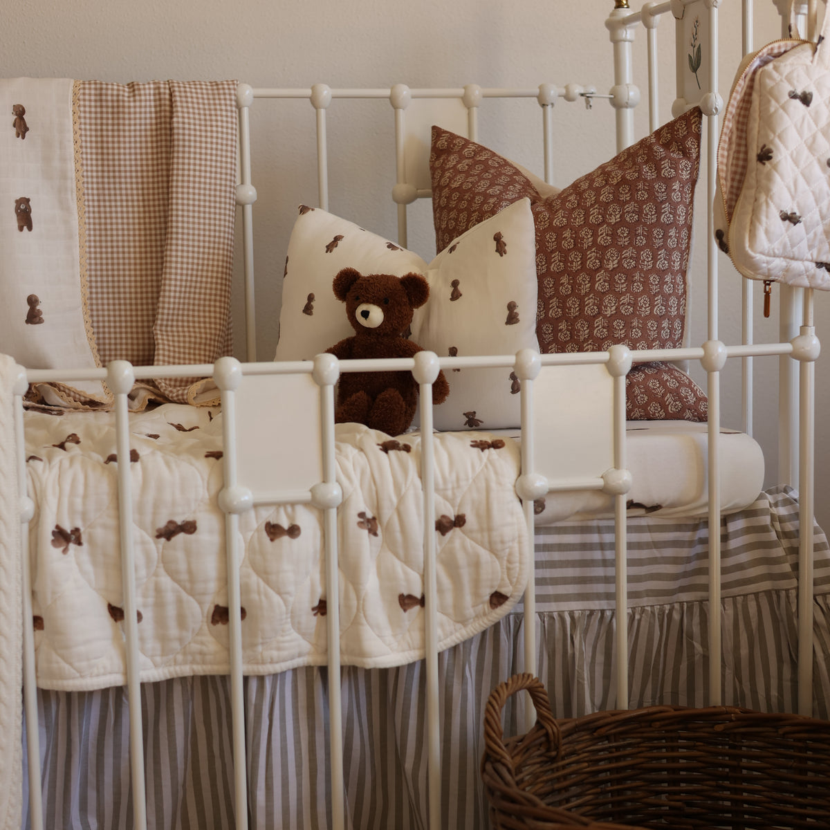 A cozy nursery crib setup with a white metal crib, featuring a cream and brown quilted bedding set with teddy bear motifs, matching pillows, a plush brown teddy bear, and a striped crib skirt.