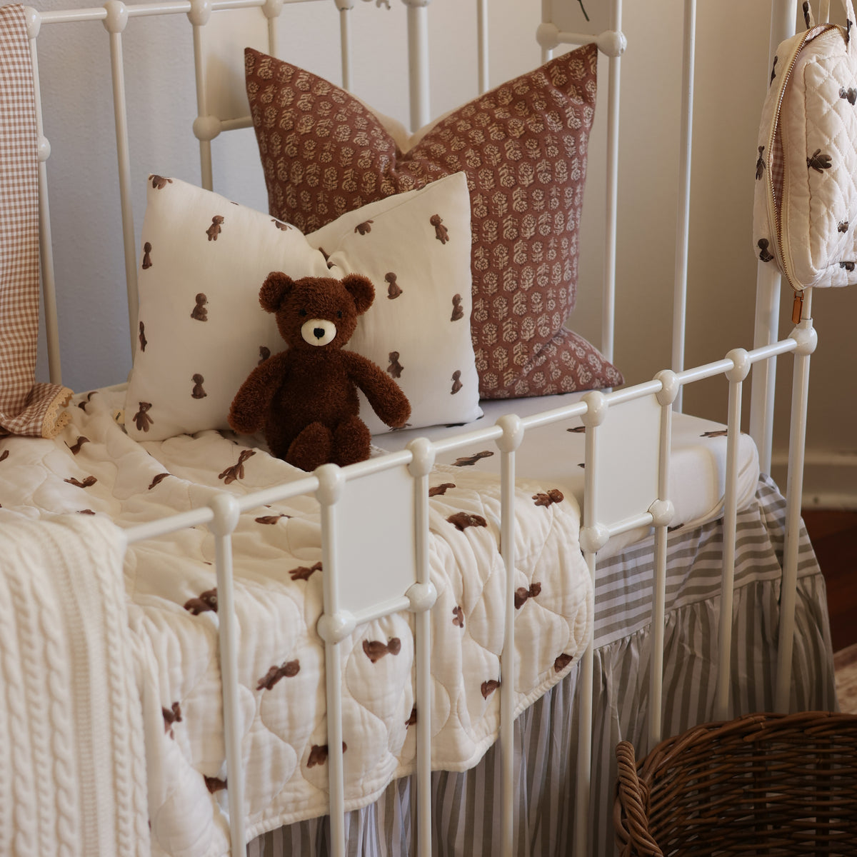 A styled nursery bed featuring a cream quilted blanket and matching pillow shams with small brown teddy bear motifs, a striped bed skirt, a brown teddy bear plush toy, and two decorative pillows in cream and brown patterned fabric, all arranged on a white metal bed frame.