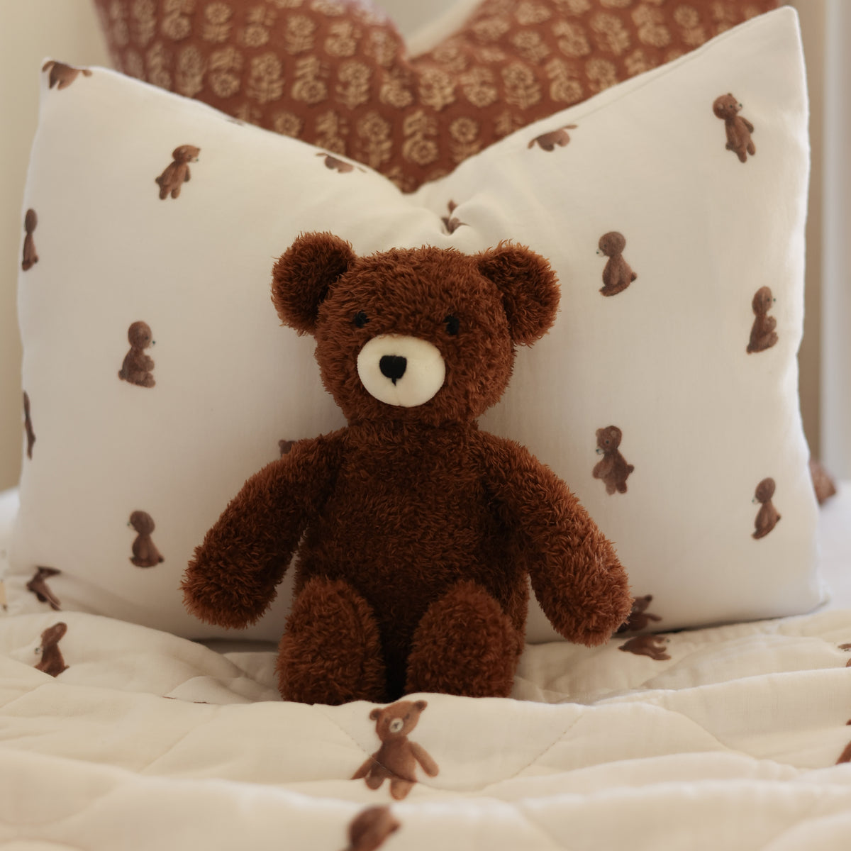 A brown plush teddy bear sits centered on a white quilted bedspread in front of two white pillows with small brown teddy bear prints. A patterned brown pillow is visible in the background.