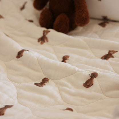 Close-up of a quilted cream or off-white blanket with small brown embroidered animal motifs, with a soft brown plush toy partially visible at the top.