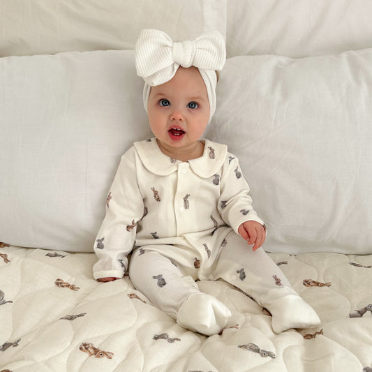 Baby in a white outfit with animal patterns sitting on a white blanket.