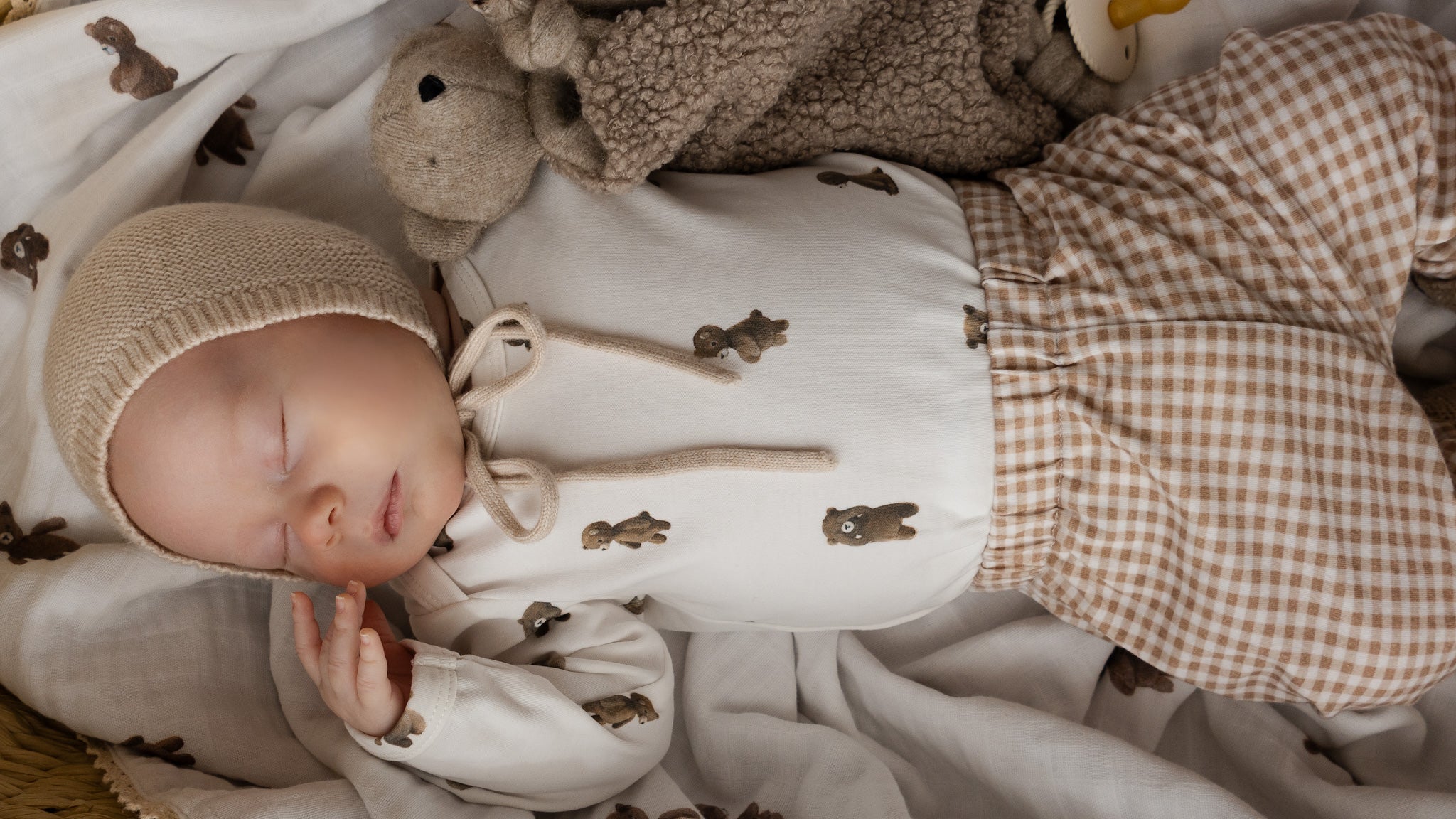 A sleeping newborn baby wearing a cream knit bonnet and matching cream bodysuit with brown rabbit prints, paired with brown and cream gingham pants, lying on a soft cream blanket with a plush teddy bear.