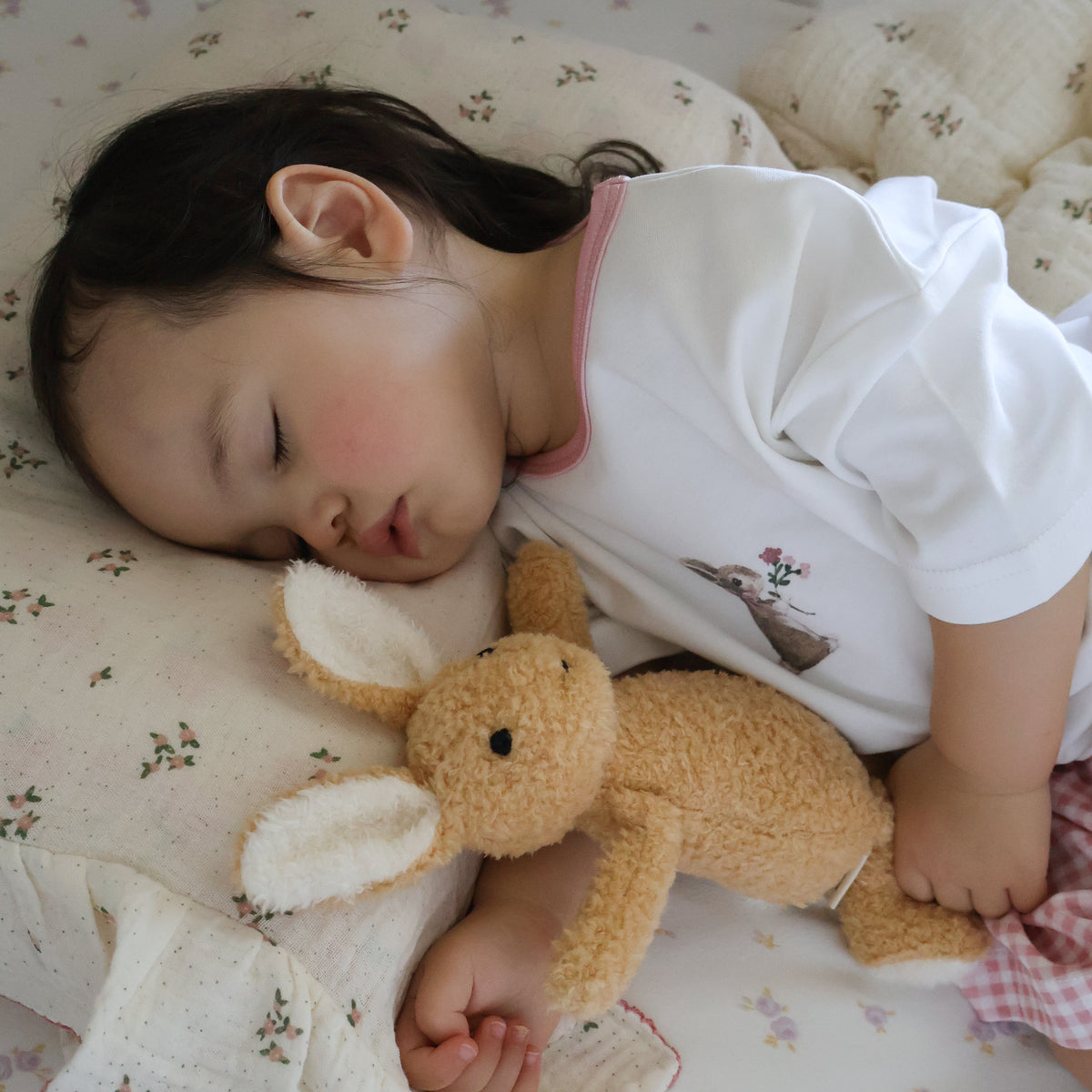 A sleeping young child lying on a floral quilt, wearing a white short-sleeve top with a pink collar and a small deer graphic on the front, while holding a plush tan bunny toy.