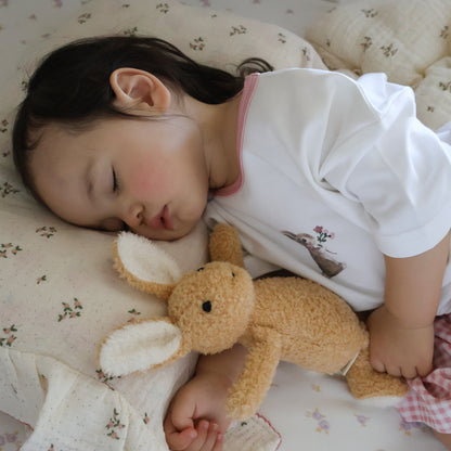 A sleeping young child lying on a floral quilt, wearing a white short-sleeve top with a pink collar and a small deer graphic on the front, while holding a plush tan bunny toy.
