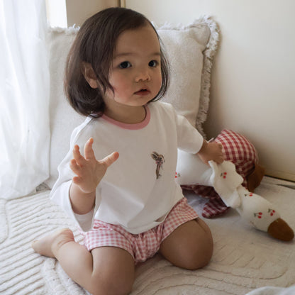 A toddler girl sitting on a cream-colored quilted bed, wearing a white long-sleeve top with a pink collar and a small graphic, paired with pink and white gingham shorts. She is holding a plush toy next to her.