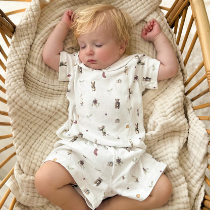 A baby sleeping peacefully in a wicker basket, wearing a short-sleeve cream cotton romper with a small woodland bear pattern, layered with a thick textured cream muslin blanket.