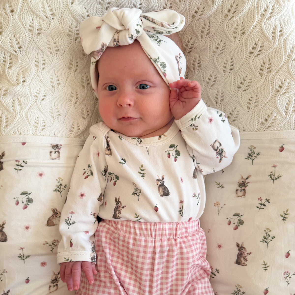 A baby lying on a textured cream blanket, wearing a cream long-sleeve top with a bunny and strawberry print, pink and white gingham pants, and a matching cream bow headband.