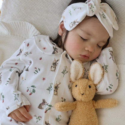 A sleeping baby wearing a white long-sleeve onesie and matching bow headband with a small bunny and floral print, lying on a cream knit blanket and holding a plush tan bunny toy.