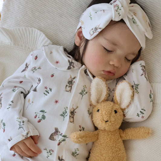 A sleeping baby wearing a white long-sleeve onesie and matching bow headband with a small bunny and floral print, lying on a cream knit blanket and holding a plush tan bunny toy.