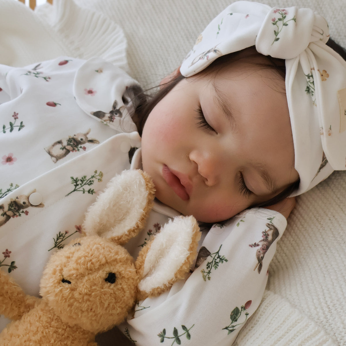 A sleeping baby wearing a white long-sleeve onesie and matching bow headband with a small bunny and floral print, resting on a cream blanket next to a plush tan bunny toy.