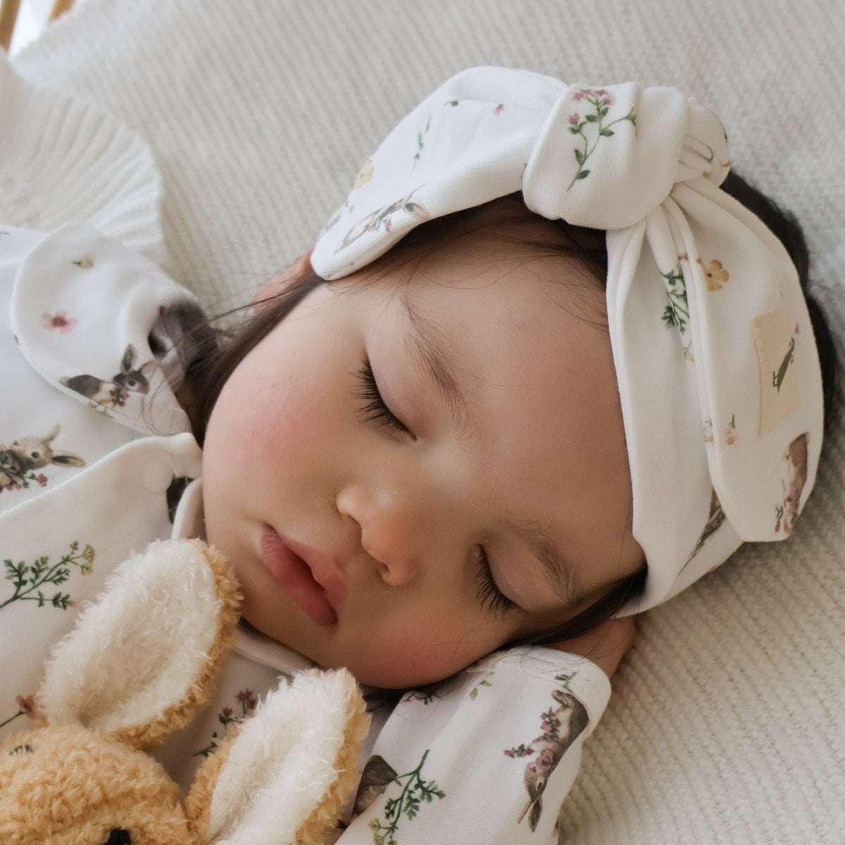 A sleeping baby wearing a matching off-white printed sleepwear set with a knotted headband and a plush bunny toy, resting on a textured cream blanket.