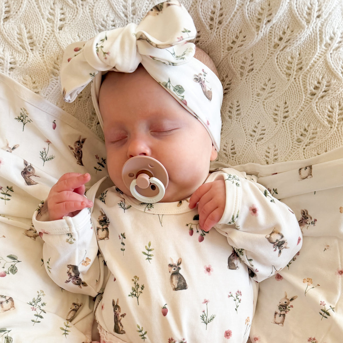 A sleeping newborn baby wearing a cream-colored long-sleeve onesie and matching bow headband, both printed with a cute bunny and floral pattern, lying on a textured cream knit blanket.
