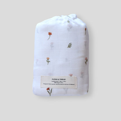 A swaddling blanket with a cream background and small floral patterns, folded neatly with a label on the front displaying the brand name.