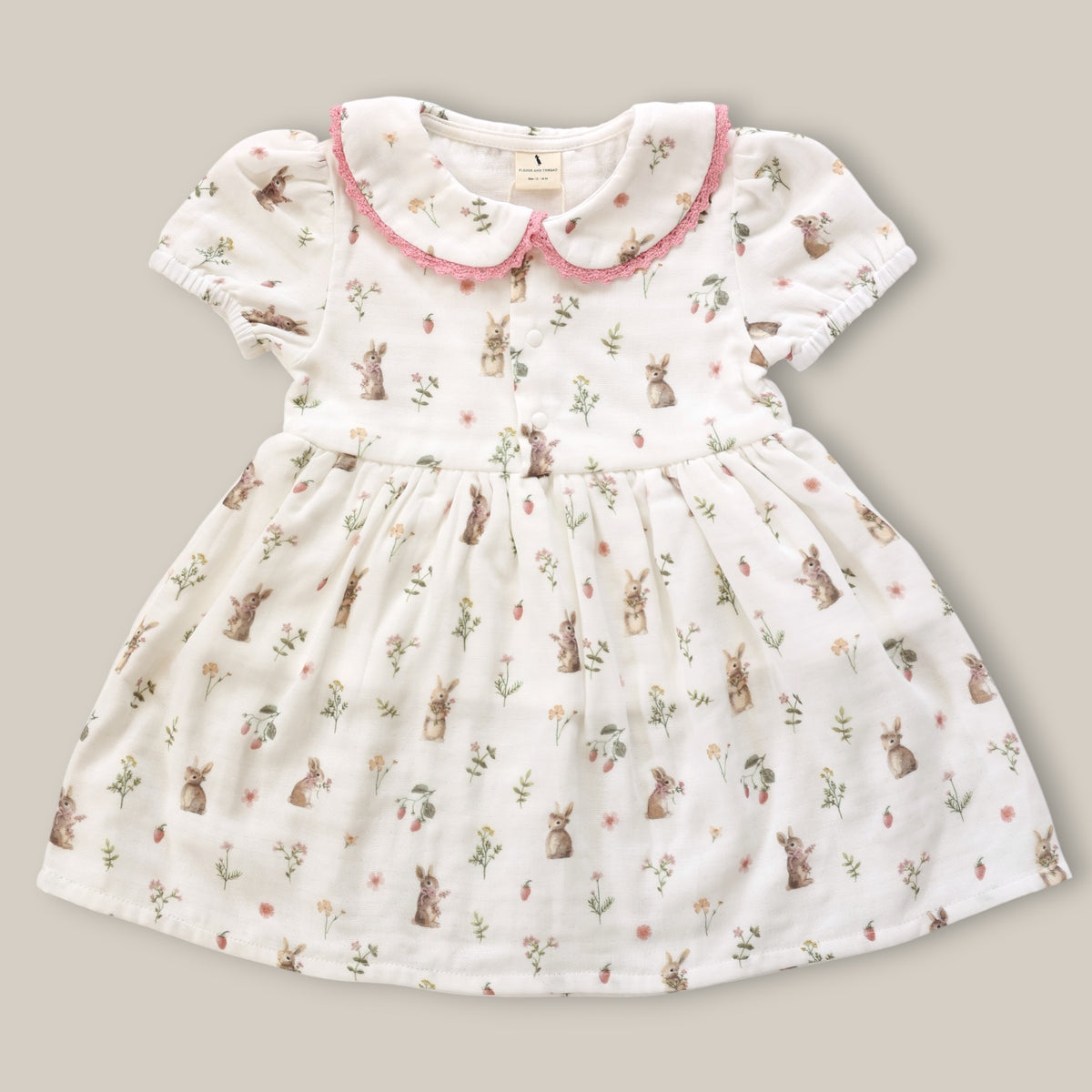 A baby girl's short-sleeve dress in white cotton muslin fabric, featuring an all-over print of brown rabbits, green leaves, and small pink flowers. The dress has a pink ruffled Peter Pan collar and a button-up front.