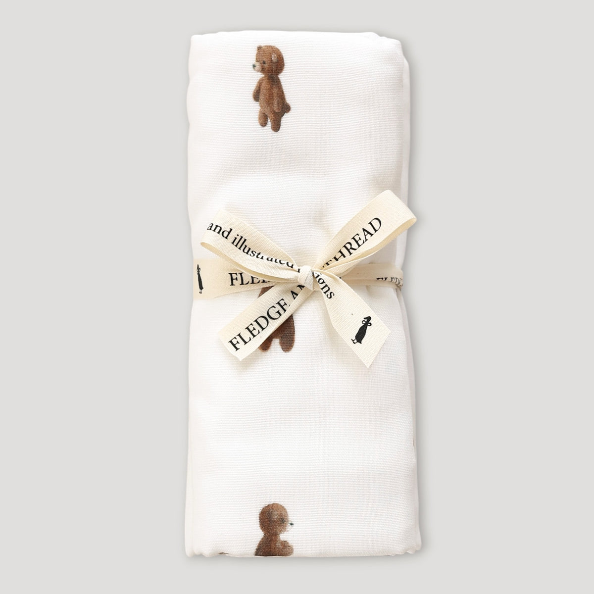 A folded white baby blanket featuring a print of small brown teddy bears, tied with a cream ribbon printed with the words 'FLEDGE MY TREAD'.