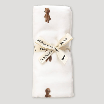 A folded white baby blanket featuring a print of small brown teddy bears, tied with a cream ribbon printed with the words 'FLEDGE MY TREAD'.