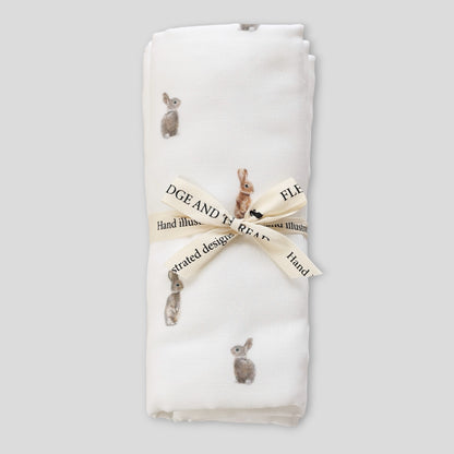 A folded white baby blanket with a print of rabbits in various poses, tied with a cream ribbon that has 'FLEDGE AND THREAD' printed on it, set against a solid light gray background.