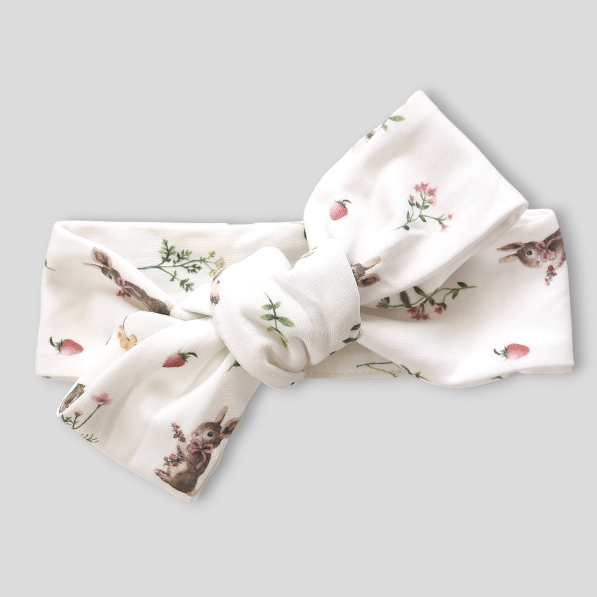 A white fabric headband with a large bow, featuring a delicate print of rabbits, strawberries, and wildflowers, shown on a solid gray background.