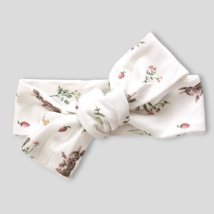 A white fabric headband with a large bow, featuring a delicate print of rabbits, strawberries, and wildflowers, shown on a solid gray background.