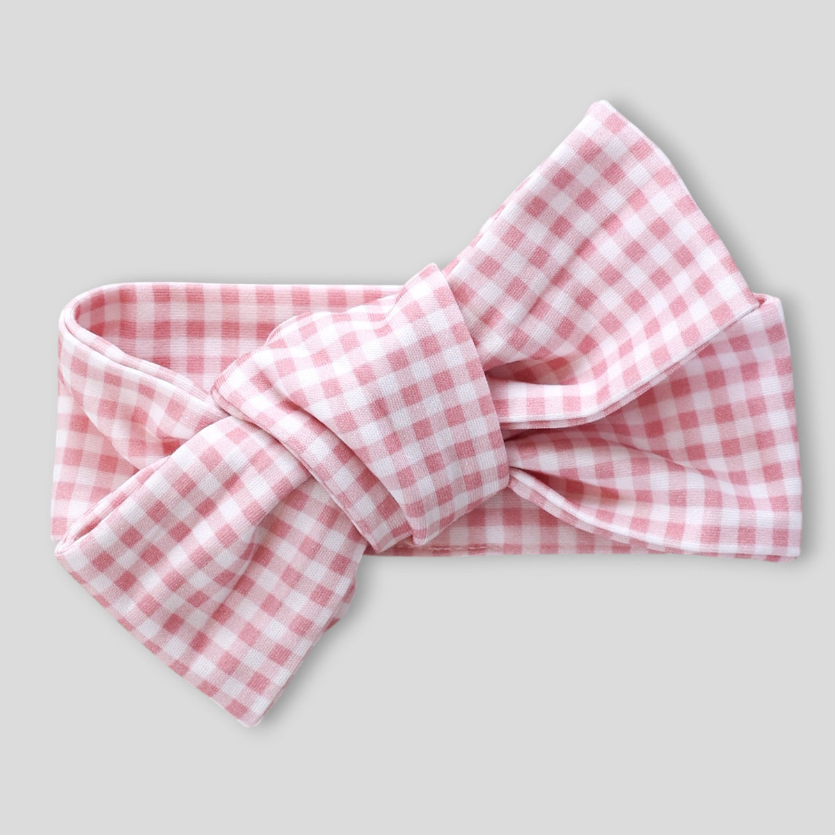 A pink and white gingham bow headband, styled with a large central knot, shown against a solid light gray background.