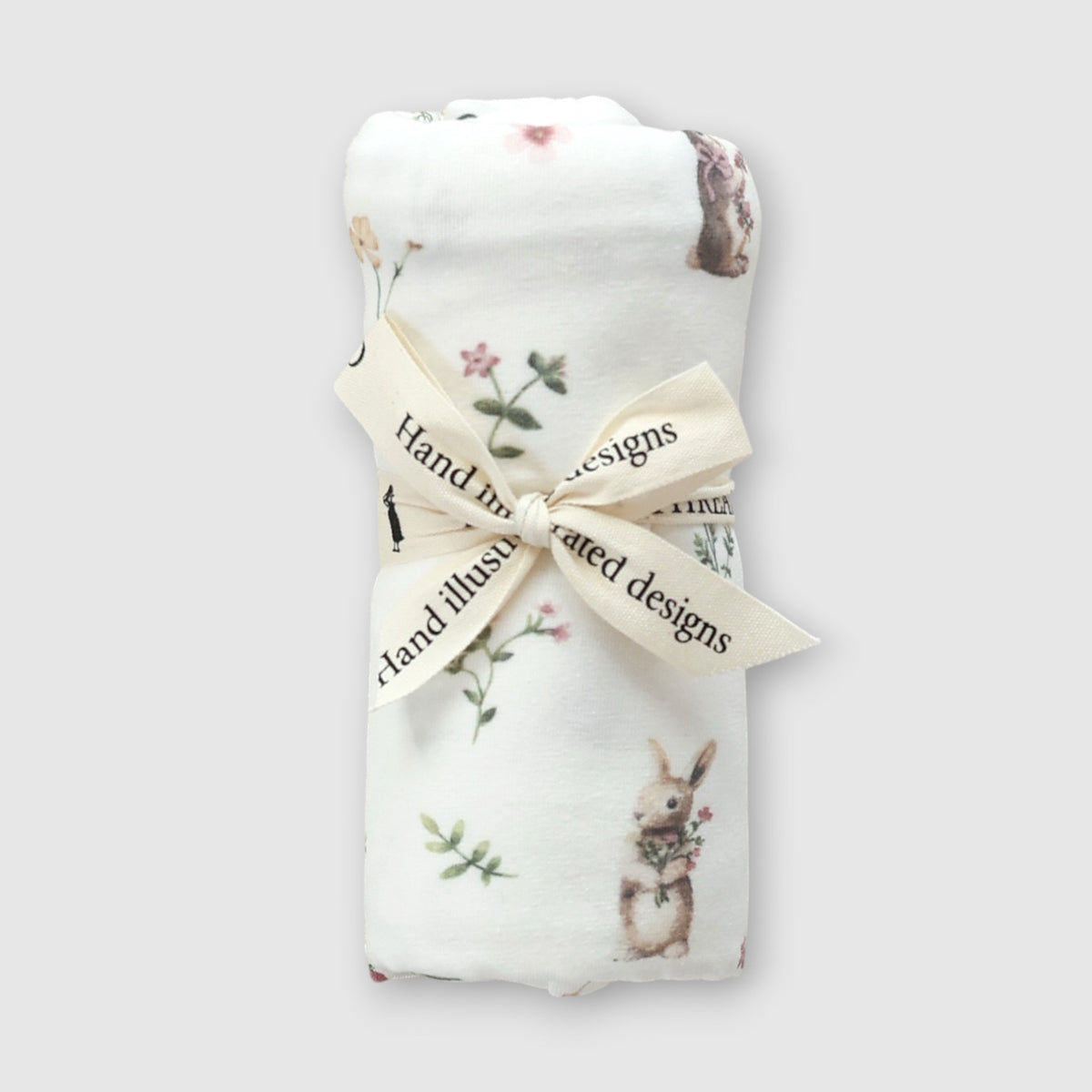 A folded white baby blanket with a printed rabbit and floral pattern, tied with a cream ribbon that reads 'Hand illustrated designs', on a light gray background.