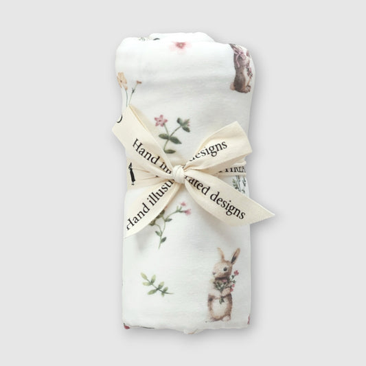A folded white baby blanket with a printed rabbit and floral pattern, tied with a cream ribbon that reads 'Hand illustrated designs', on a light gray background.