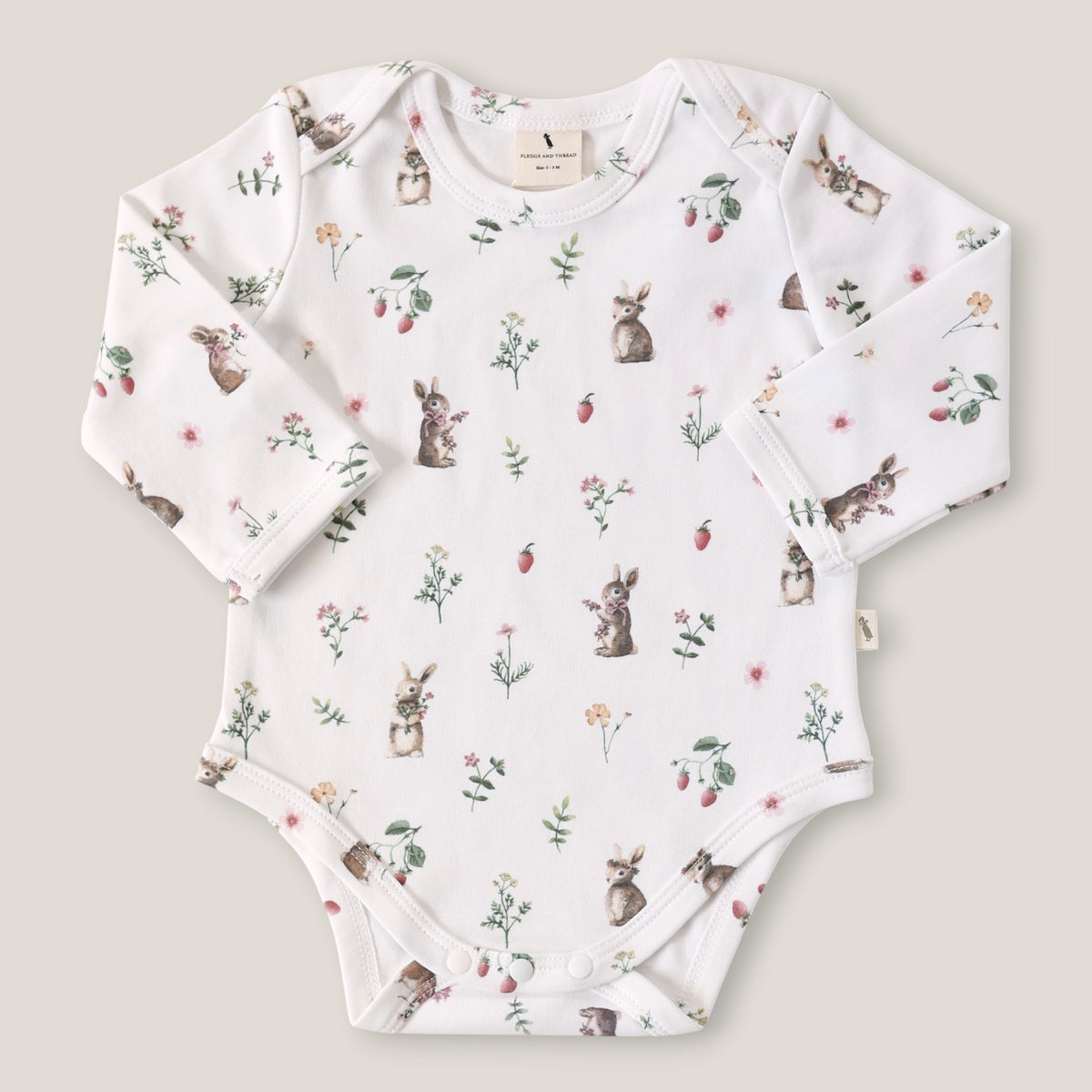 A long-sleeve baby bodysuit in white, featuring an all-over print of rabbits, strawberries, and wildflowers. The bodysuit is laid flat on a light beige background.