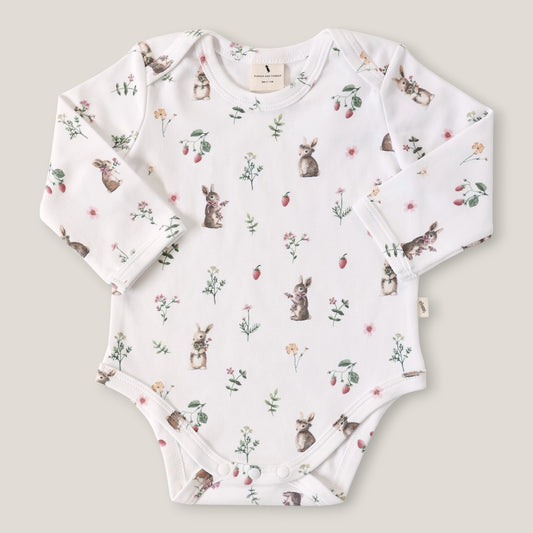 A long-sleeve baby bodysuit in white, featuring an all-over print of rabbits, strawberries, and wildflowers. The bodysuit is laid flat on a light beige background.