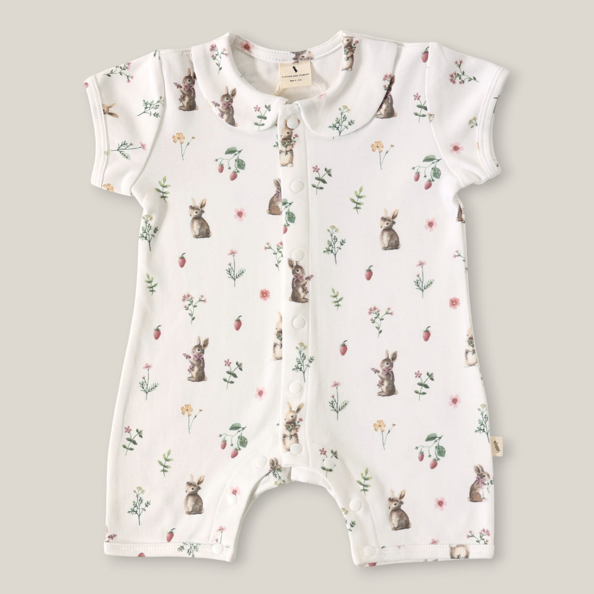 A baby short-sleeve romper in white with an all-over print of rabbits, strawberries, and small wildflowers, laid flat on a neutral beige background.