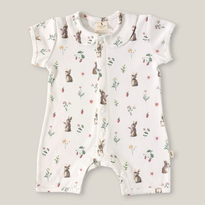 A baby short-sleeve romper in white with an all-over print of rabbits, strawberries, and small wildflowers, laid flat on a neutral beige background.
