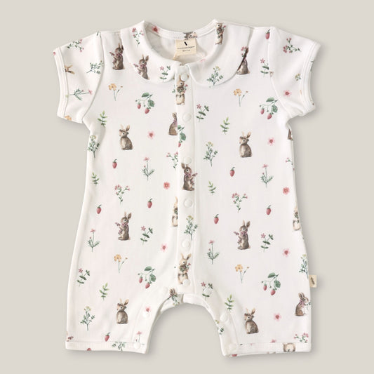 A baby short-sleeve romper in white with an all-over print of rabbits, strawberries, and small wildflowers, laid flat on a neutral beige background.