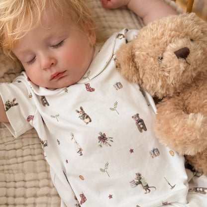 A sleeping baby wearing a white short-sleeve bodysuit with a cute teddy bear and bunny print, lying next to a plush brown teddy bear on a textured beige blanket.