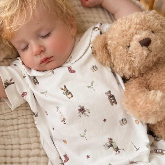 A sleeping baby wearing a white short-sleeve bodysuit with a cute teddy bear and bunny print, lying next to a plush brown teddy bear on a textured beige blanket.